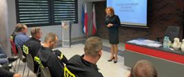 W dolnej części zdjęcia jeden rząd strażaków, na środku prowadząca w mundurze, za nią wyświetlana prezentacja. W rogu sali ustawione flagi Polski Po prawej biurko. 