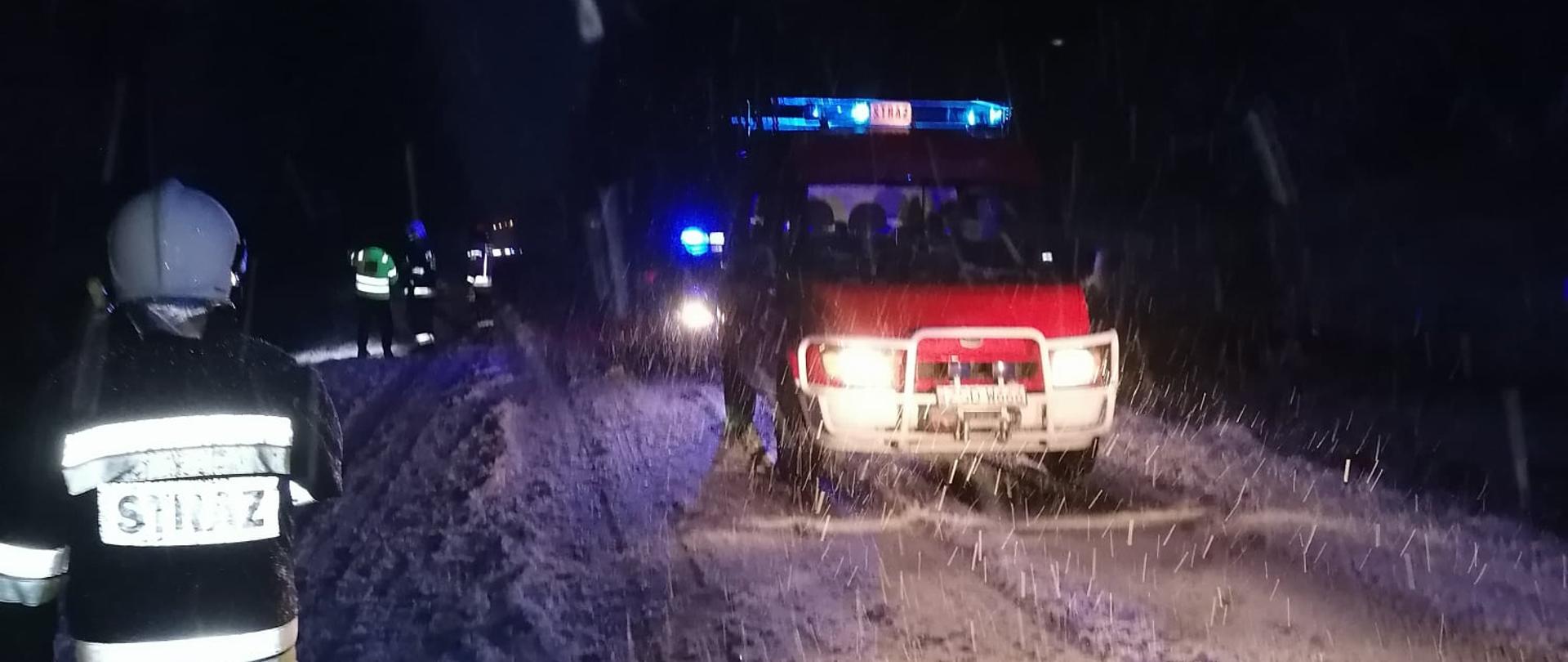 Zdjęcie przedstawia zaśnieżoną drogę. Na pierwszym planie widzimy strażaka w ubraniu specjalnym oraz białym hełmie oraz pojazd strażacki z włączonymi światłami mijania oraz niebieskimi błyskowymi. Za pojazdem strażackim widać kolejny pojazd uprzywilejowany z włączonymi ww. światłami prawdopodobnie jest to pojazd Policji oraz po jego lewej stronie osoby z odblaskami.