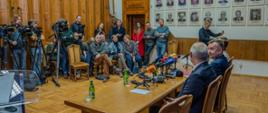 Minister Wieczorek i dwóch mężczyzn w garniturach siedzą za drewnianym stołem, przed nimi kilkunastu dziennikarzy.