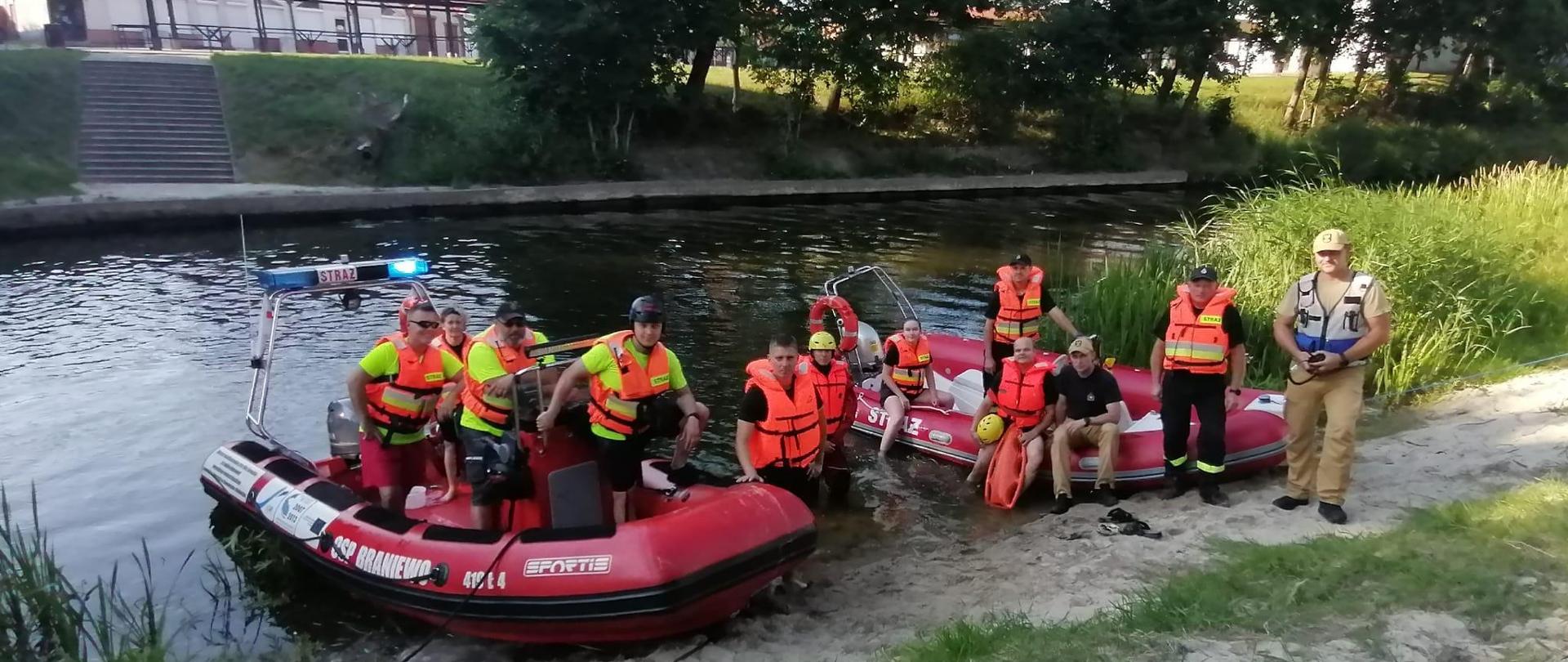 Nas brzegu rzeki stoją dwie łodzie strażackie, na łodziach i brzegu strażacy w pomarańczowych kamizelkach. Słonecznie. W tle rzeka i drugi brzeg.