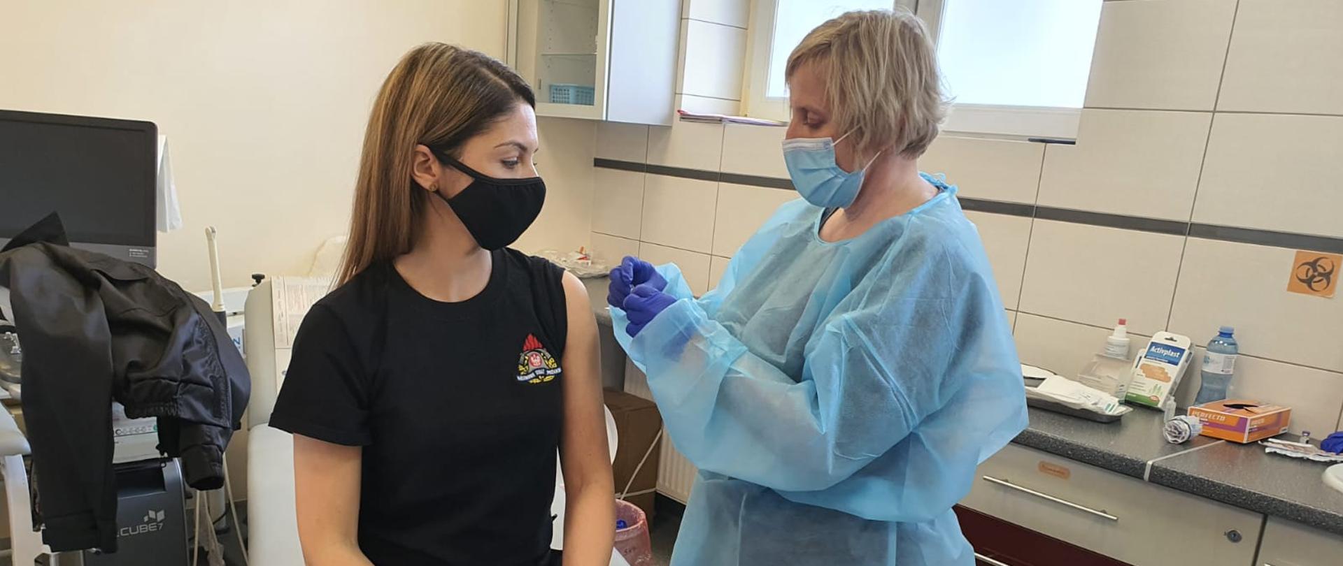 Na zdjęciu strażak (kobieta) ubrana w ubranie koszarowe oraz koszulkę koloru czarnego siedzi na łóżku w gabinecie lekarskim. Po prawej stronie pielęgniarka ubrana w fartuch podaje szczepionkę w lewe ramię strażaka. Osoby zabezpieczone w maseczki jednorazowe.