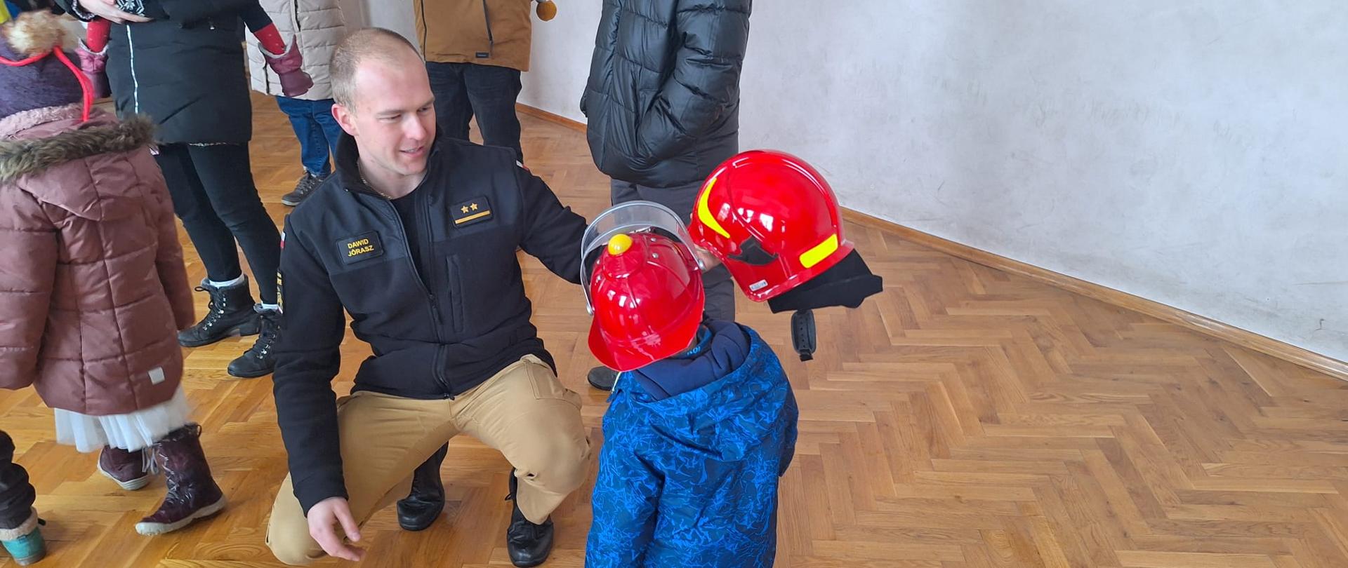 Strażak prezentuje dziecku hełm strażacki, po lewej stoją zgromadzeni goście.