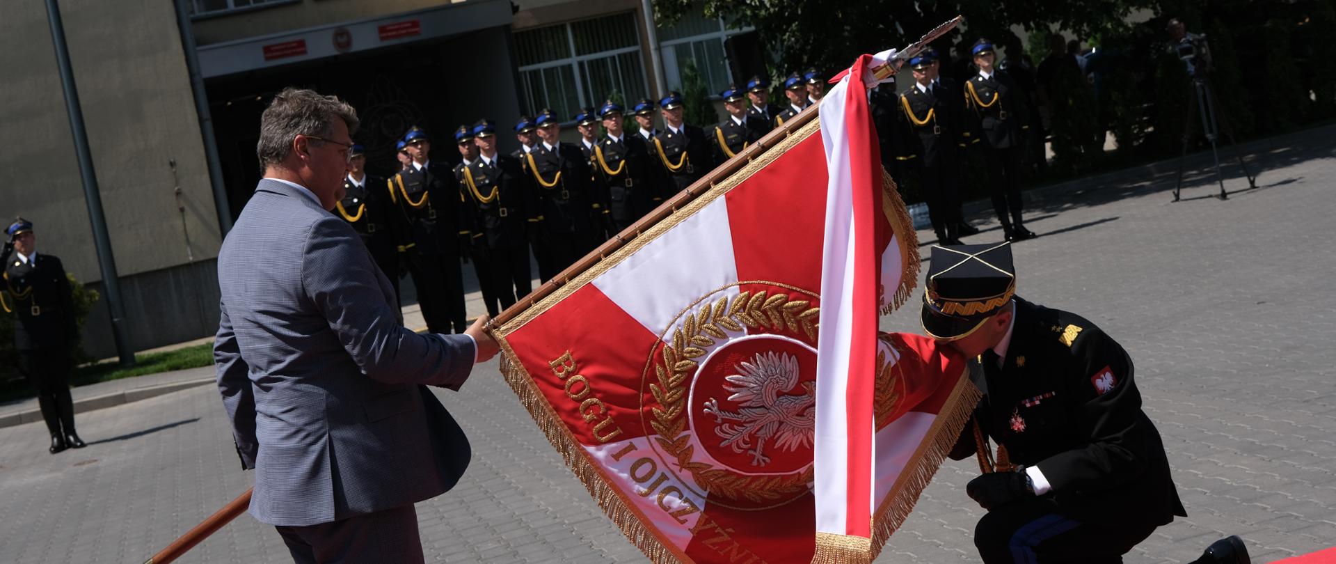 Wiceminister spraw wewn. i admin. trzyma sztandar pochylony do przodu, przed sztandarem klęka komendant główny PSP całując róg sztandaru