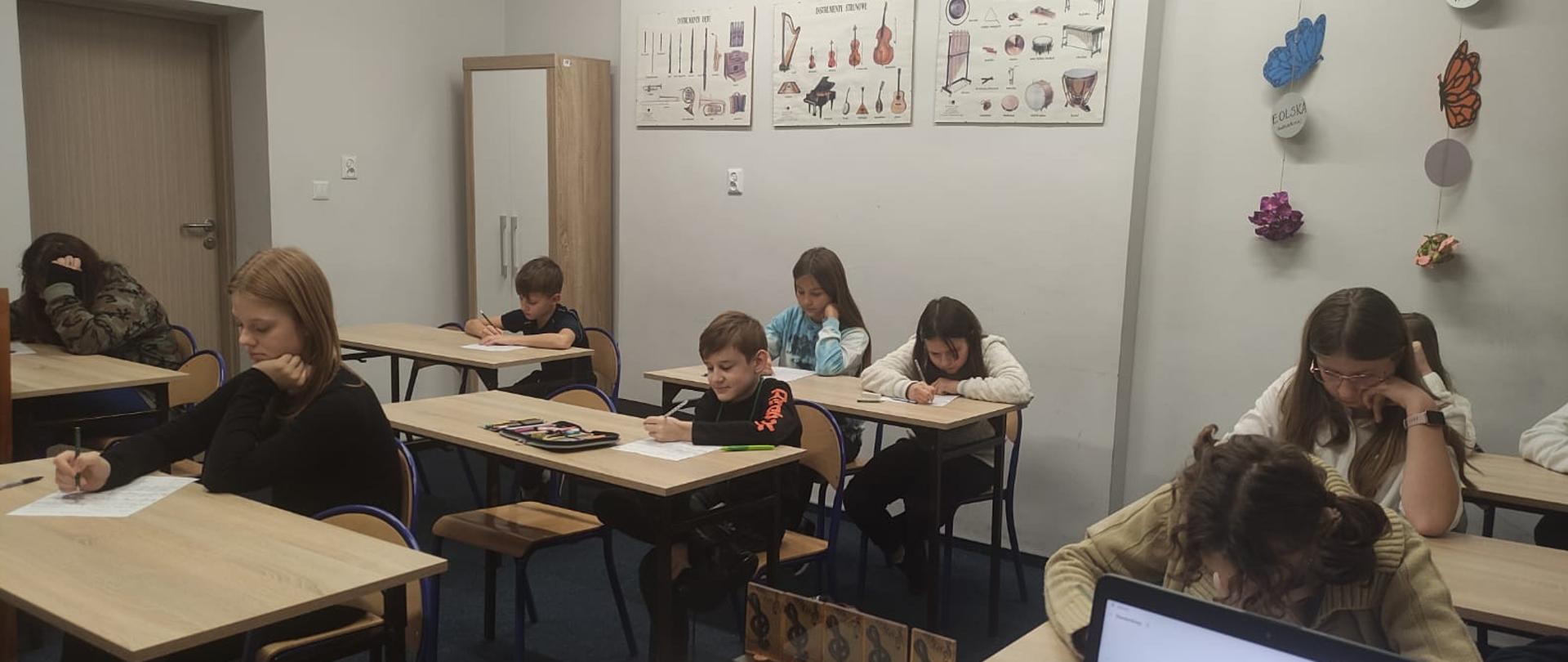 Zdjęcie przedstawia uczniów siedzących w ławkach i rozwiązujących test mający wyłonić laureatów I Szkolnego Konkursu ,, Mistrz Dyktanda", w tle ilustracje przedstawiające instrumenty. 