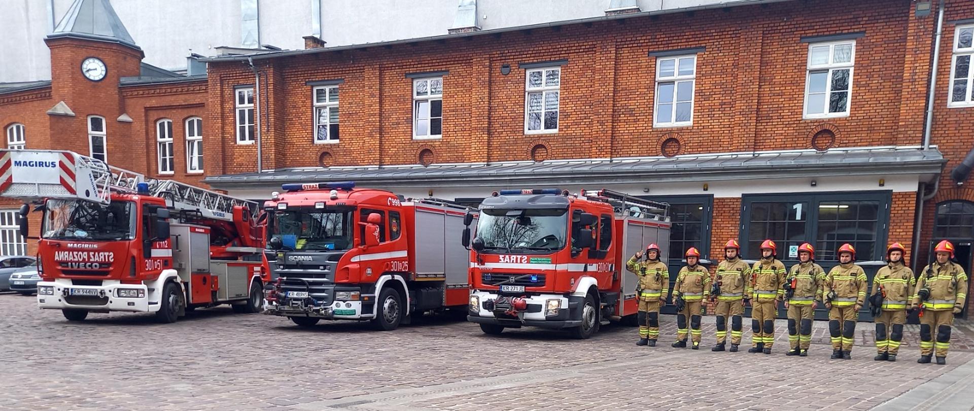 Strażacy stoją w szeregu na placu obok czerwonych samochodów ratowniczo-gaśniczych