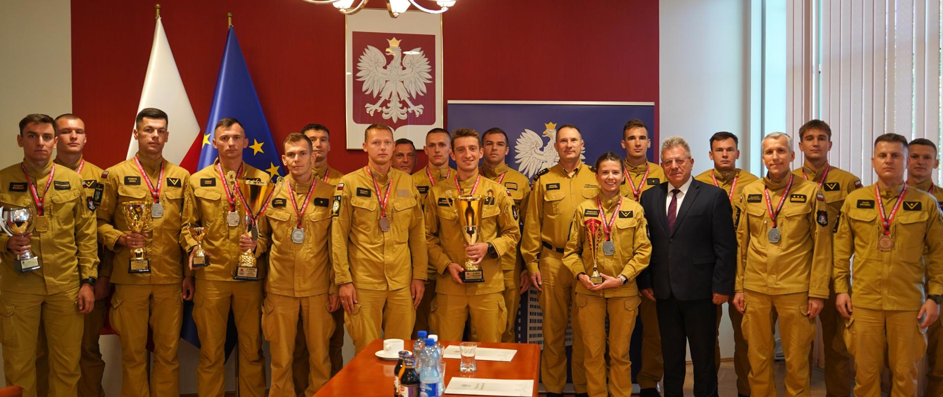 Pamiątkowe zdjęcie grupowe strażaków Państwowej Straży Pożarnej z medalami i pucharami oraz cywilnego oficjela w sali konferencyjnej pod godłem Rzeczypospolitej Polskiej.