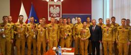 Pamiątkowe zdjęcie grupowe strażaków Państwowej Straży Pożarnej z medalami i pucharami oraz cywilnego oficjela w sali konferencyjnej pod godłem Rzeczypospolitej Polskiej.