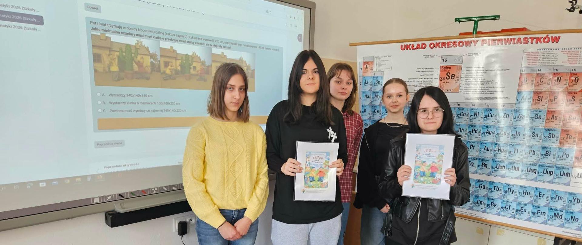 Grupa uczennic stojących przy tablicy multimedialnej, dwie trzymają dyplomy