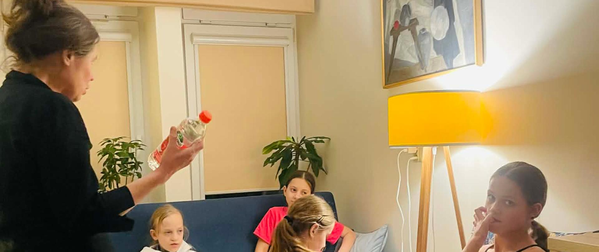 Zdjęcie czterech dziewczynek w pokoju wychowawców razem z wychowawczynią. Jedna z dziewczynek siedzi na fotelu, dwie siedzą na kanapie a czwarta klęczy obok lampy. Wychowawczyni trzyma w ręku butelkę pepsi.