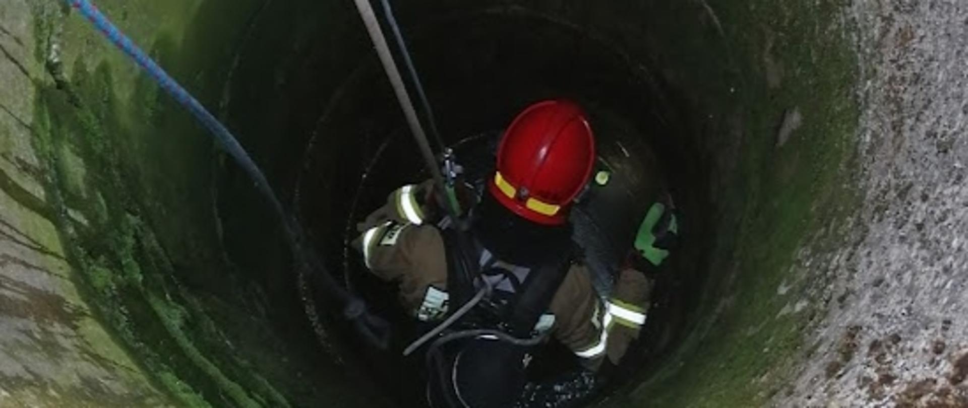 Strażak wyposażony w sprzęt ochrony układu oddechowego, w szelkach alpinistycznych, podczepiony do lin, podczas zejścia do wnętrza studni.