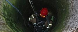 Strażak wyposażony w sprzęt ochrony układu oddechowego, w szelkach alpinistycznych, podczepiony do lin, podczas zejścia do wnętrza studni.