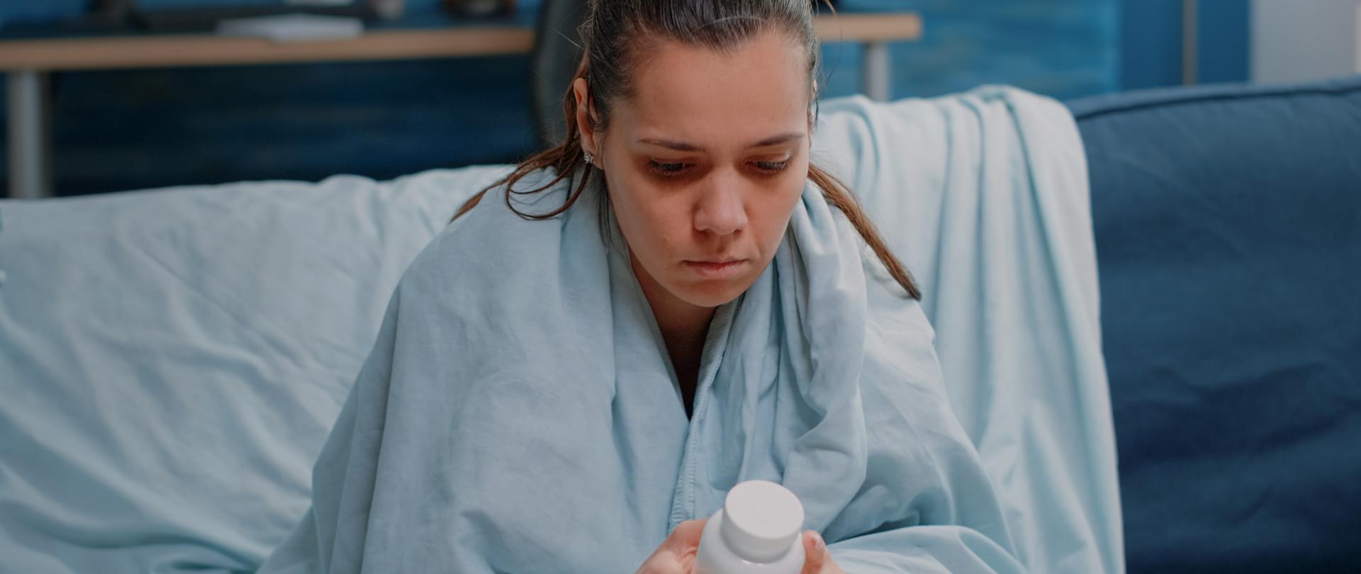Sick woman with flu holding bottle of pills and drugs, reading label of medication to cure coronavirus symptoms. Person looking at flask of medicament for prescription treatment.