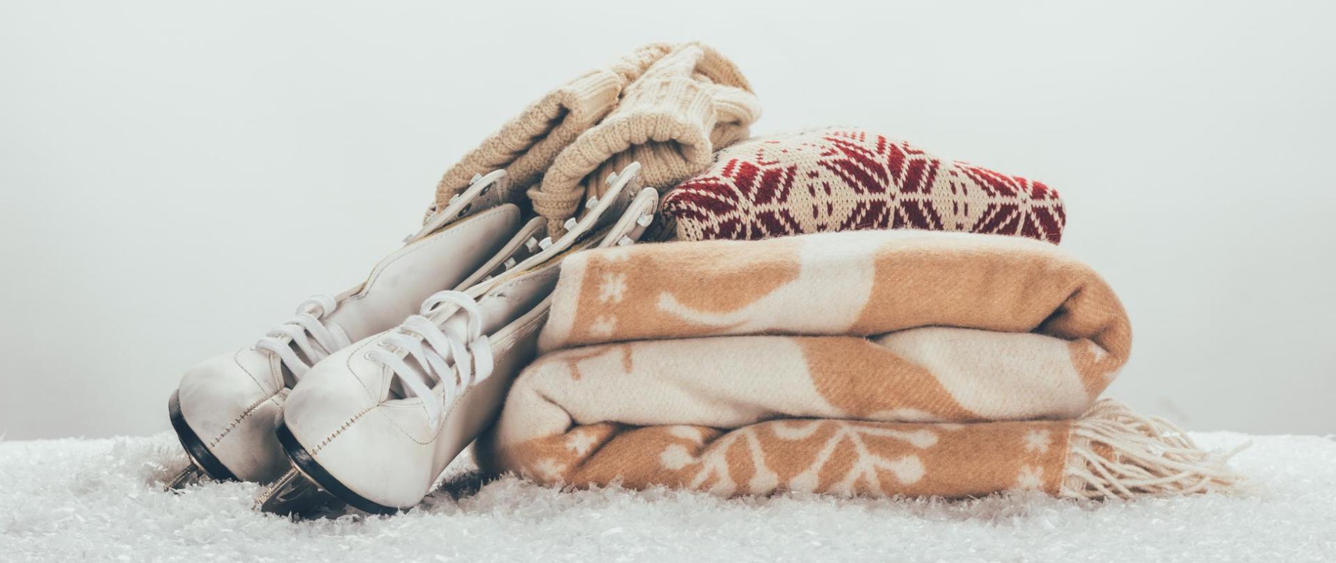 Na zdjęciu są łyżwy oparte o złożony koc i sweter. Ponadto na łyżwach leżą rękawiczki. Przedmioty leżą na dywanie.