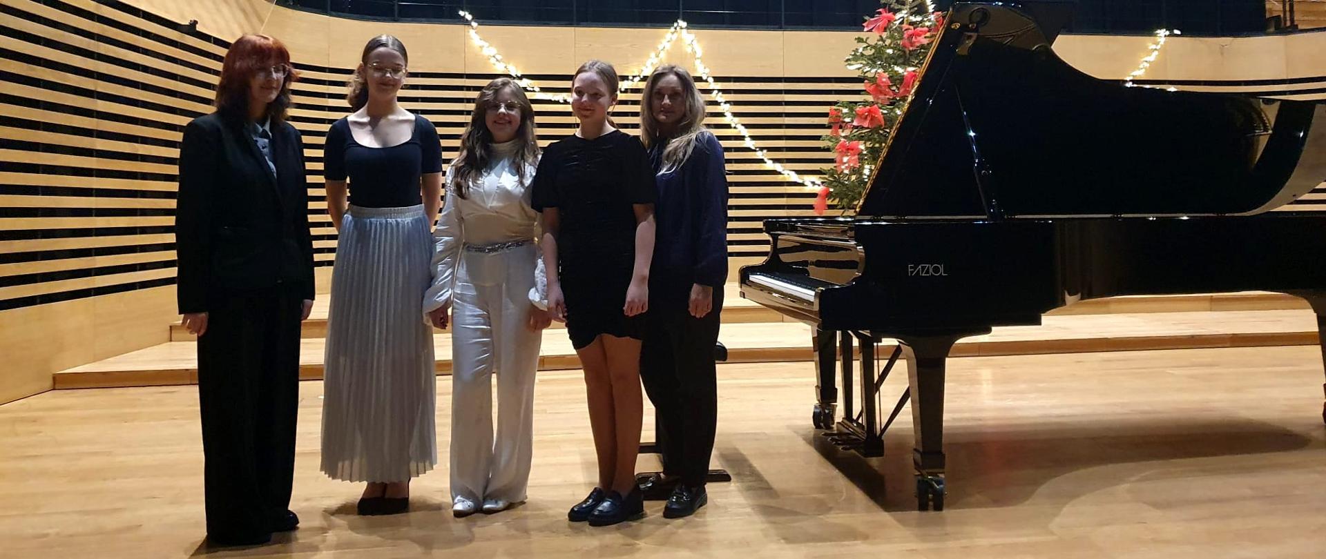 Kobieta i czworo dziewcząt stoi w jednym rzędzie na estradzie sali koncertowej, obok pop prawej stronie stoi fortepian.
