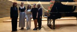 Kobieta i czworo dziewcząt stoi w jednym rzędzie na estradzie sali koncertowej, obok pop prawej stronie stoi fortepian.