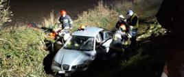 Samochód osobowy marki Seat wjechał do rowu. Przy nim znajduje się trzech strażaków oraz policjant którzy wyciągają poszkodowane osoby. 