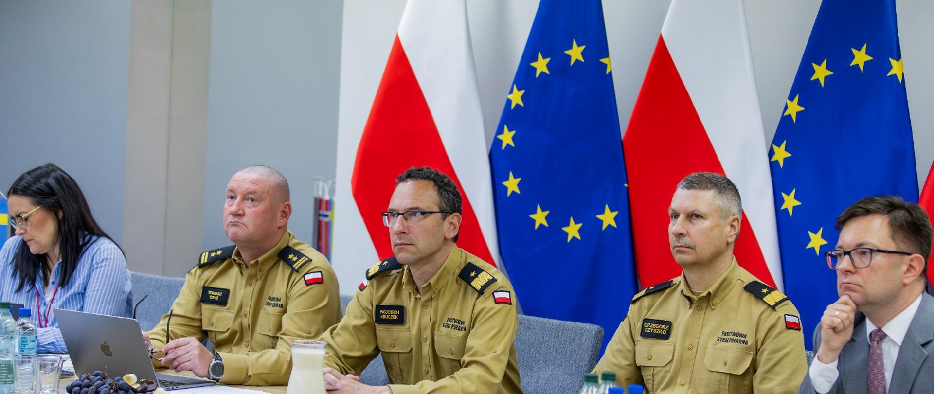 Grupa osób, w tym trzech mężczyzn w mundurach koloru żółtego Państwowej Straży Pożarnej oraz dwóch cywilów, siedzi przy stole konferencyjnym z laptopami i dokumentami; w tle widoczne są flagi Polski i Unii Europejskiej.