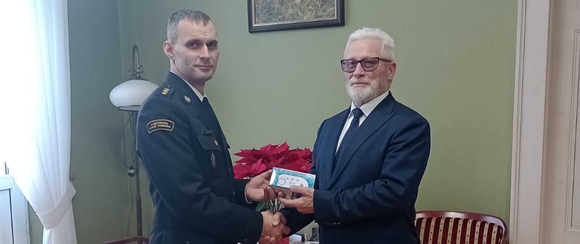 KOLEJNY ROK POWIAT FUNDUJE CZUJKI DLA MIESZKAŃCÓW. Na zdjęciu Starosta Kościański wręcza symbolicznie czujkę tlenku węgla na ręce Komendanta Powiatowego PSP w Kościanie.