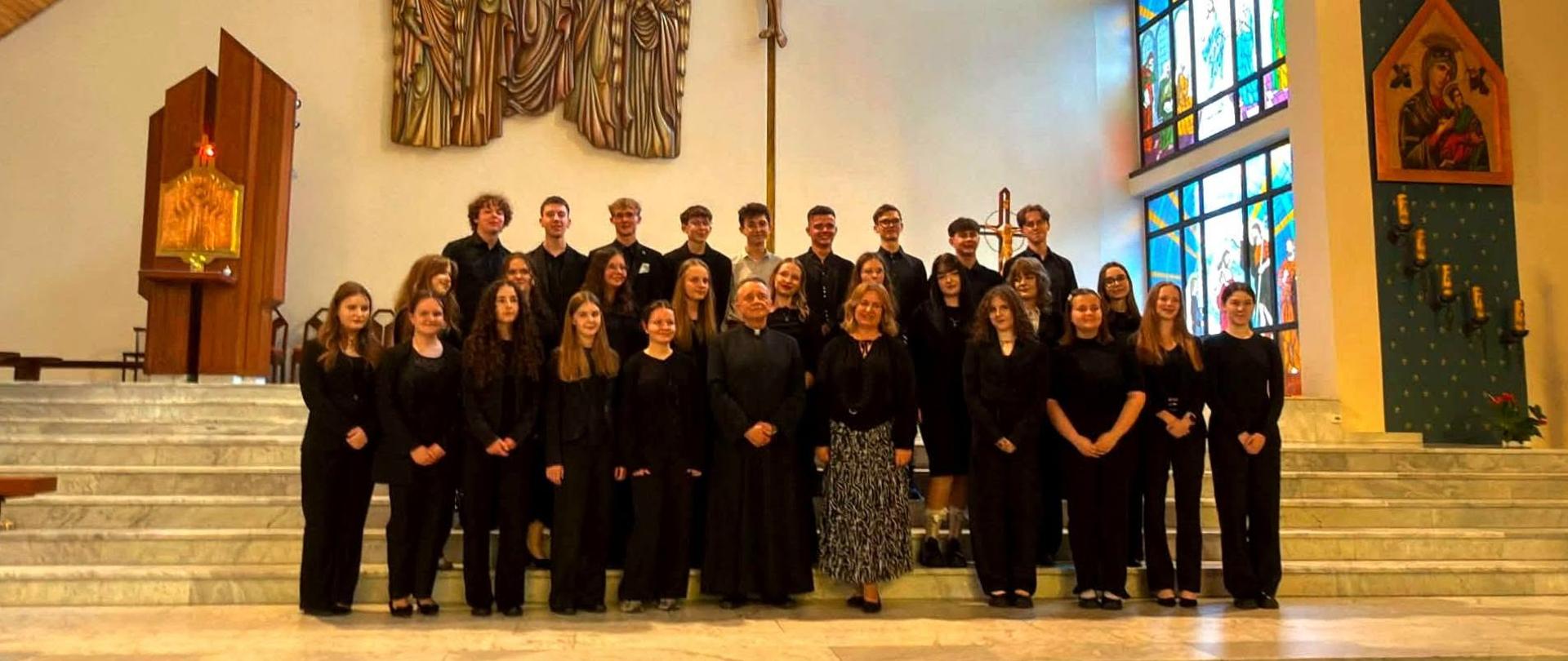 Zdjęcie grupowe chóru w kościele. Około dwudziestu pięciu osób ubranych w czarne stroje stoi na schodach prowadzących do ołtarza. W tle widoczne są charakterystyczne elementy wnętrza kościoła: duża metalowa rzeźba przedstawiająca scenę religijną na lewej ścianie, kolorowy witraż z geometrycznymi wzorami po prawej stronie, drewniana boazeria na skośnym suficie oraz ikona religijna w niszy po prawej stronie. Oświetlenie jest ciepłe, a marmurowe schody i posadzka tworzą eleganckie tło dla grupy. 