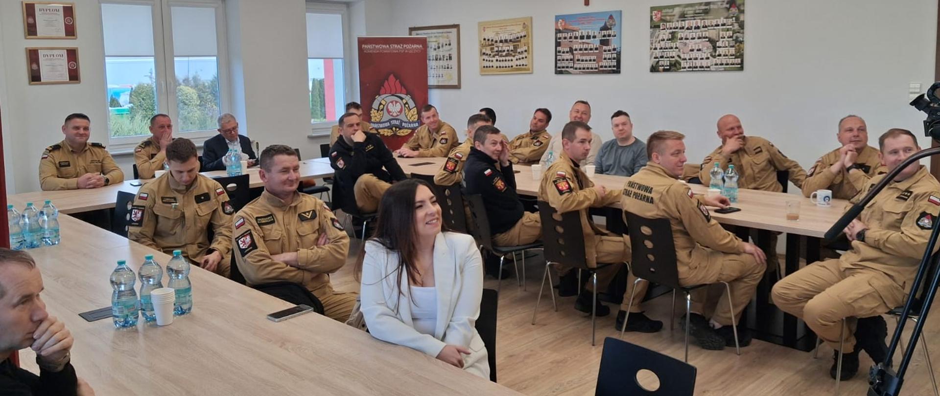Na zdjęciu widać uczestników szkolenia – głównie strażaków w umundurowaniu, którzy siedzą przy stołach w sali konferencyjnej. Na ścianach widoczne są dyplomy oraz tablo ze zdjęciami. 