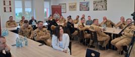Na zdjęciu widać uczestników szkolenia – głównie strażaków w umundurowaniu, którzy siedzą przy stołach w sali konferencyjnej. Na ścianach widoczne są dyplomy oraz tablo ze zdjęciami. 