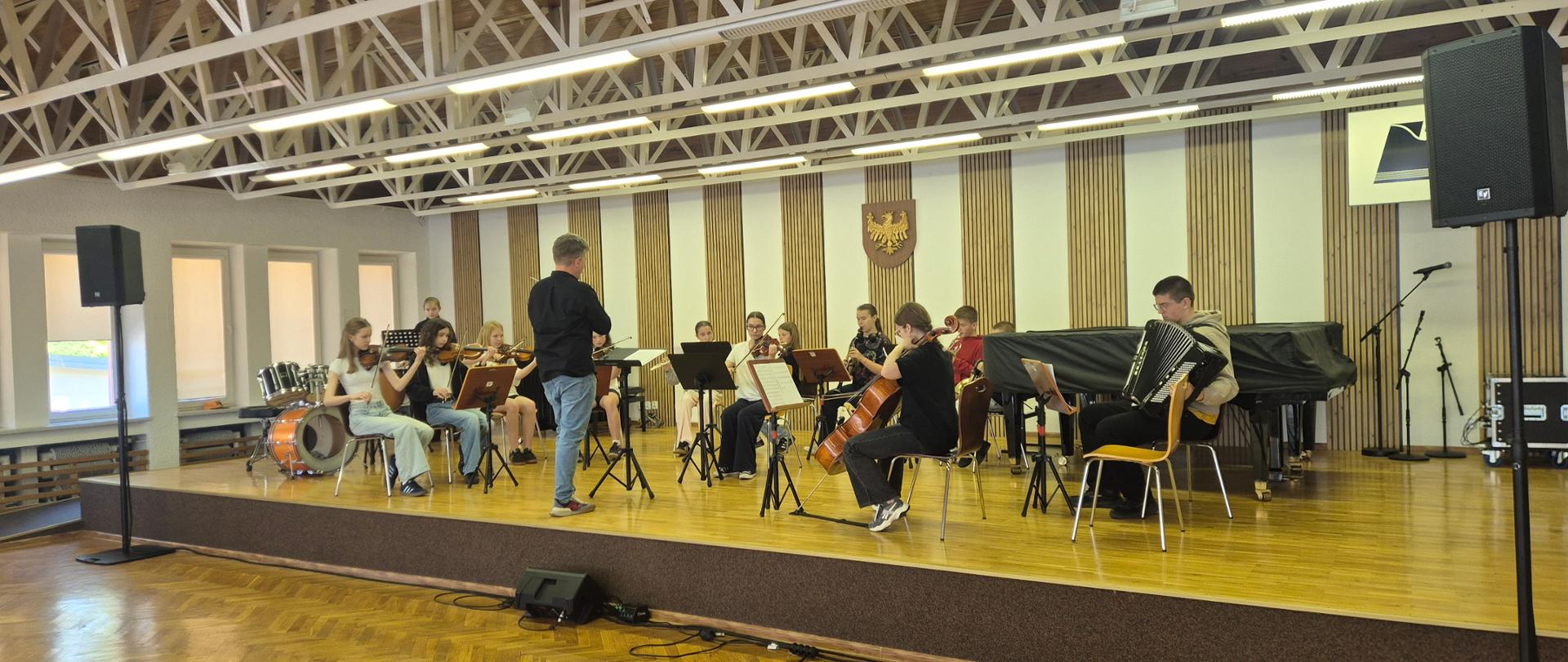  Na zdjęciu widać orkiestrę szkolną złożonych z uczniów grających na instrumentach smyczkowych oraz dętych. Przewodzi im dyrygujący nauczyciel.
