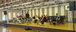  Na zdjęciu widać orkiestrę szkolną złożonych z uczniów grających na instrumentach smyczkowych oraz dętych. Przewodzi im dyrygujący nauczyciel.