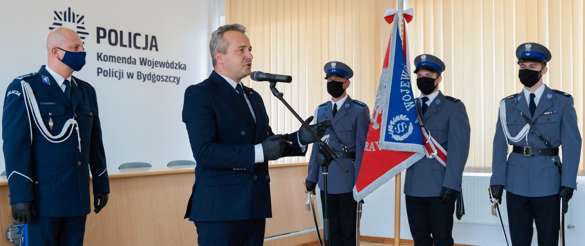 Przemawia wojewoda Mikołaj Bogdanowicz, na drugim planie sztandar Komendy Wojewódzkiej Policji oraz Komendant Wojewódzki insp. Piotr Leciejewski