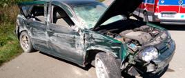Zdjęcie przedstawia samochód po wypadku - zielony VW Golf. w Tle widać pojazd pogotowia ratunkowego oraz pole.