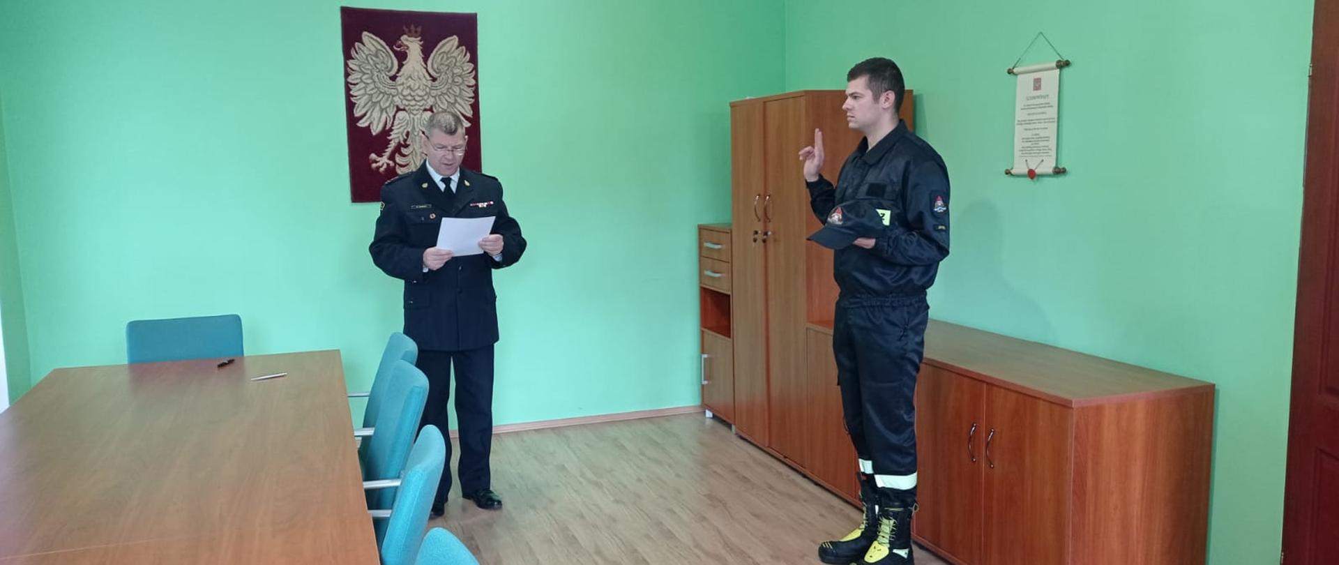 Obraz przedstawia śrubowanie nowego strażaka. Komendant Powiatowy odczytuje słowa roty. Strażacy w sali odpraw.