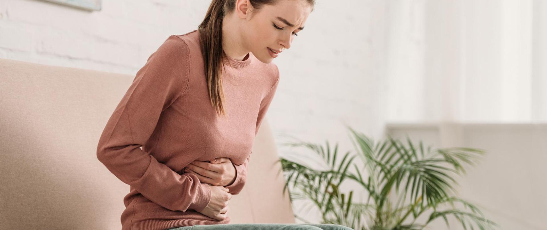 Na zdjęciu jest kobieta siedząca na kanapie oraz trzymająca się za brzuch.