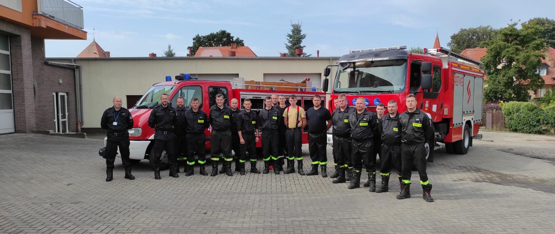 Na zdjęciu widać grupę strażaków w w umundurowaniu służbowym. W tle dwa pojazdy pożarnicze