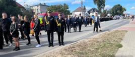 Dwóch funkcjonariuszy Państwowej Straży Pożarnej stoi przed miejscem wypadku gdzie położone są znicze za nimi stoją uczestnicy uroczystości pogrzebowej.