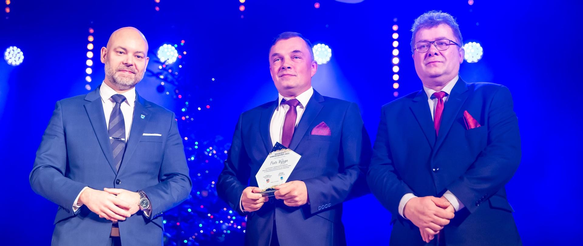 Trzech mężczyzn w garniturach stoi na scenie Samorządowego Centrum Kultury w Mielcu; jeden z nich trzyma statuetkę otrzymaną za szczególne zasługi w dziedzinie kultury.