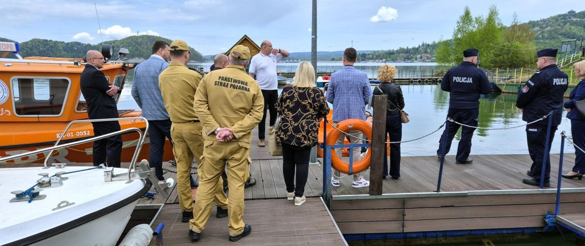 Zdjęcie przedstawia komendanta PSP z Ustrzyk Dolnych, zastępcę komendanta PSP z Leska oraz przedstawicieli władz samorządowych i innych służb którzy stoją nad jeziorem Solińskim na przystani WOPRu. Na zdjęciu widać również przycumowaną łódż WOPRu. 