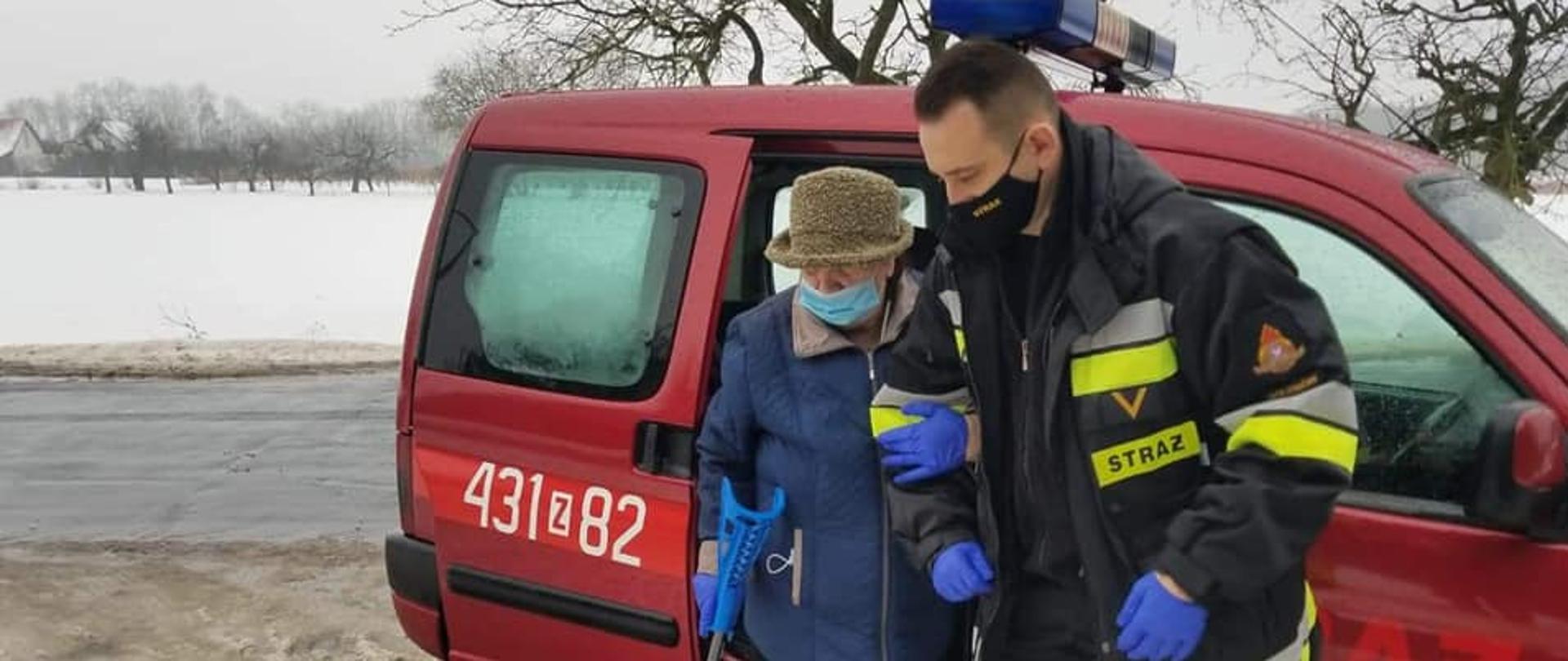 Funkcjonariusz PSP prowadzący po rękę przewożoną osobę starszą po przewiezieniu do punktu szczepień, znajdują się zaraz przy samochodzie służbowym. W tle drzewo znajdujące się przy drodze, za drzewem znajduje się pole pokryte śniegiem. 