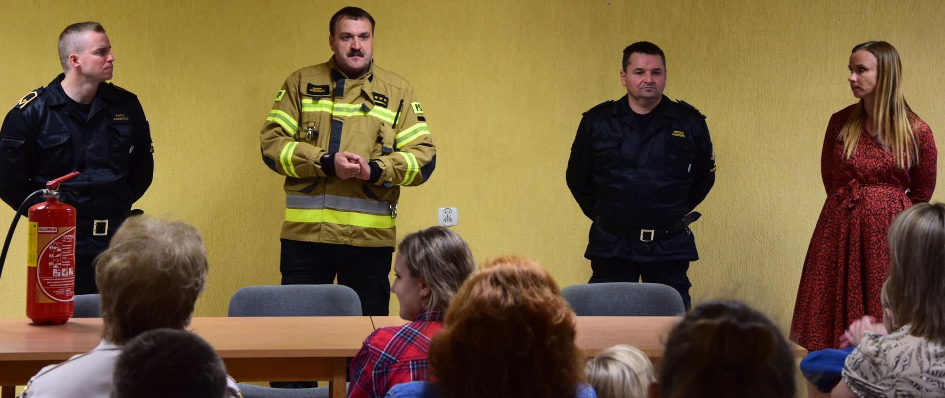 Trzech mężczyzn strażaków oraz kobieta stoją pod ścianą, od osób siedzących na krzesłach przed nimi oddzielają ich drewniane stoliki, na których stoi gaśnica.