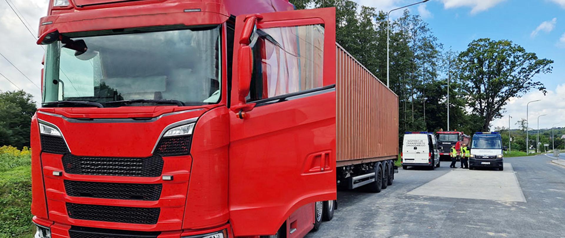 Czerwony ciągnik siodłowy z otwartymi drzwiami kabiny, ciągnący naczepę kontenerową, zaparkowany na poboczu drogi. W tle widoczne są pojazdy Inspekcji Transportu Drogowego oraz funkcjonariusze prowadzący kontrolę.