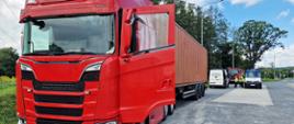 Czerwony ciągnik siodłowy z otwartymi drzwiami kabiny, ciągnący naczepę kontenerową, zaparkowany na poboczu drogi. W tle widoczne są pojazdy Inspekcji Transportu Drogowego oraz funkcjonariusze prowadzący kontrolę.