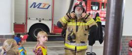 Na zdjęciu widać strażaka w mundurze bojowym obok którego widać dzieci z przedszkola. Wszyscy znajdują się w garażu jednostki. W tle widać samochód strażacki. 