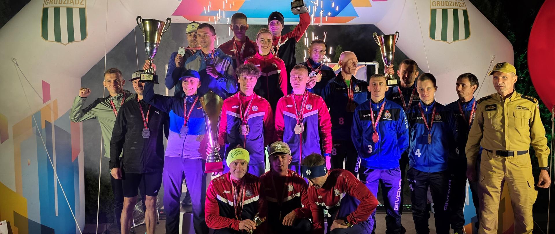 Podium biegu w nocnej porze. Na poszczególnych stopniach stoi 3 drużyny. Po bokach dwóch strażaków.