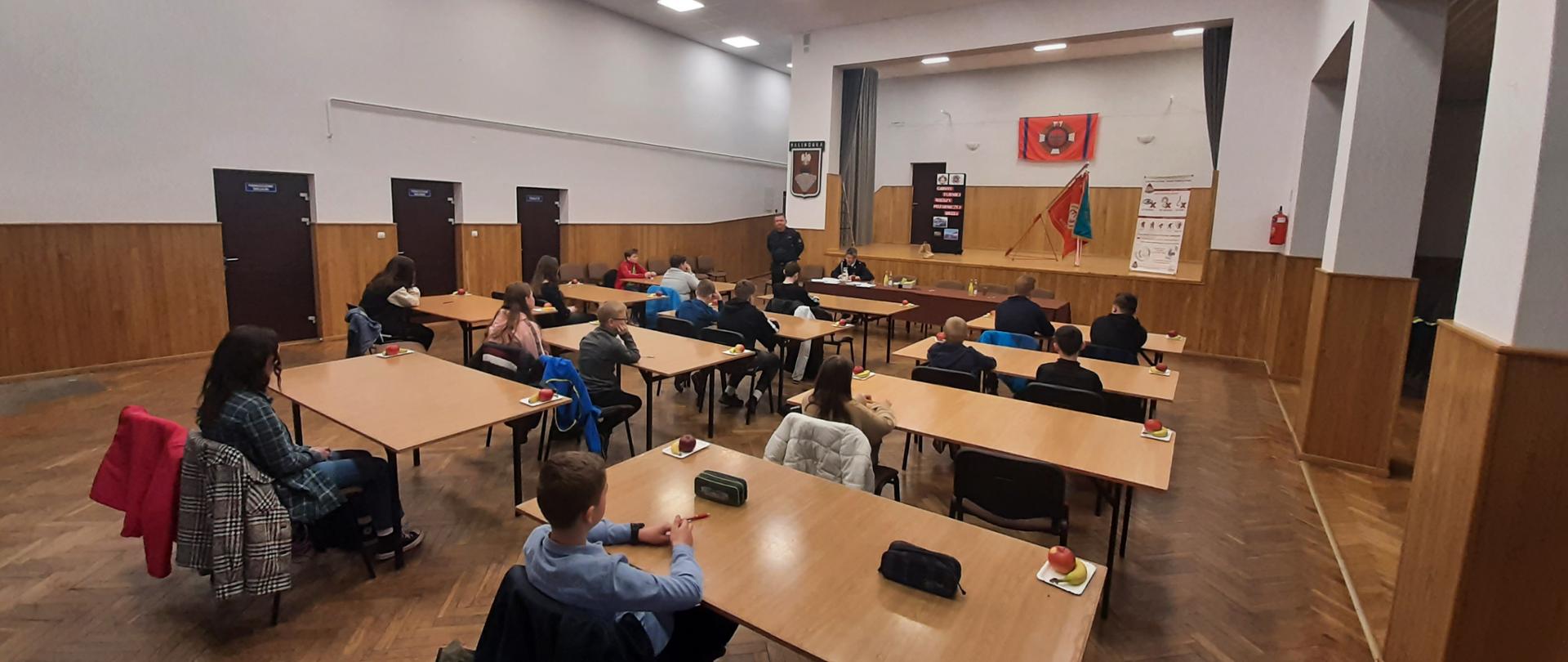 Kolorowa fotografia wykonana wewnątrz pomieszczenia. Przedstawia ona dużą jasną salę, na której stoją stoliki i krzesła. Przy nich siedzi kilkanaście młodych osób. Na końcu sali stół i dwóch umundurowanych strażaków. W tle scena ze sztandarem i banerami informacyjnymi.