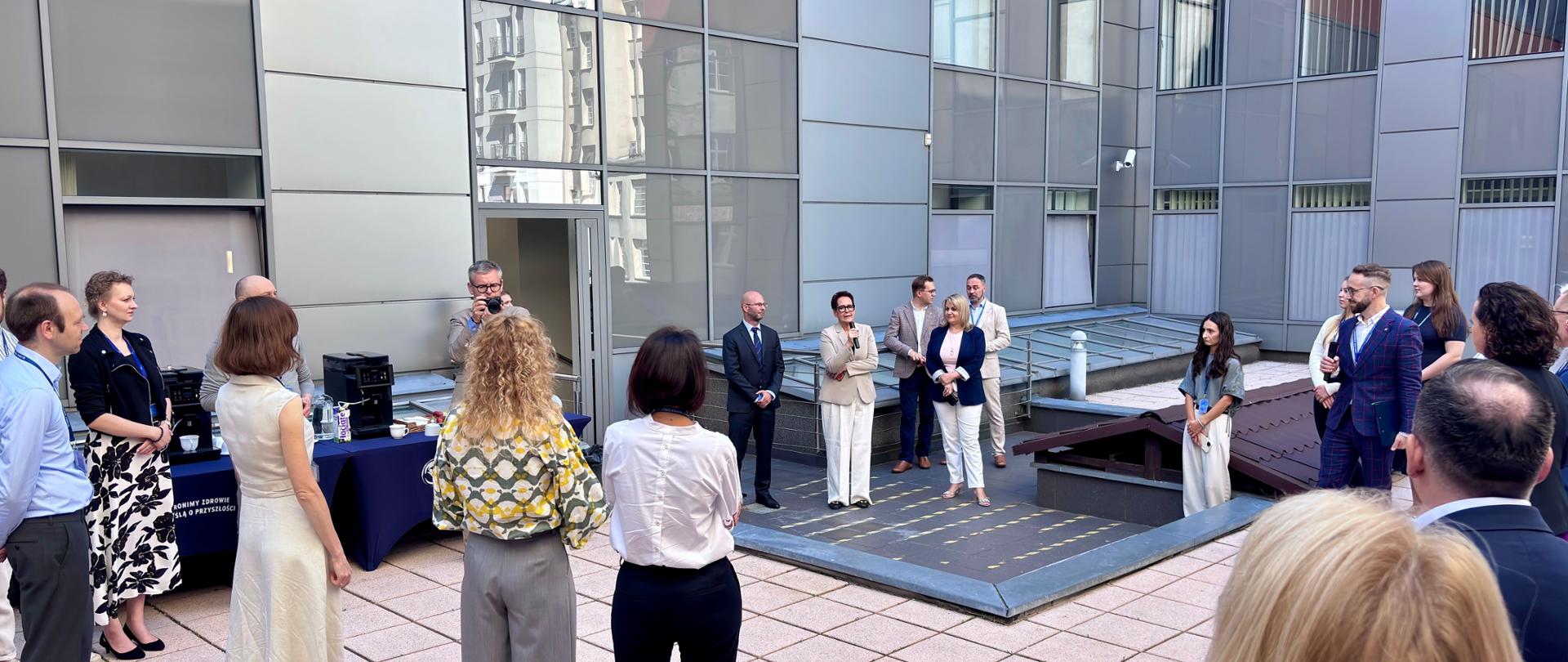 Szefowa służby cywilnej przemawia do mikrofonu na patio w Głównym Inspektoracie Sanitarnym, kilkanascie ludzi wokoł