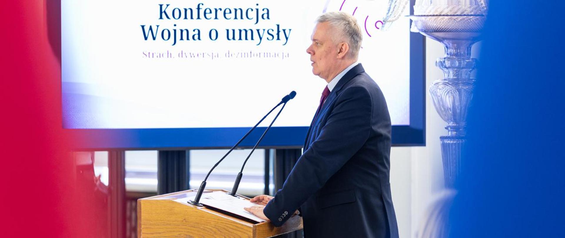 Na zdjęciu minister Tomasz Siemoniak podczas swojego wystąpienia w Sejmie na konferencji pt. "Wojna o umysły. Strach, dywersja, dezinformacja". Minister stoi za drewnianą mównicą z dwoma czarnymi mikrofonami kierunkowymi. Na mównicy teczka z materiałami. W tle monitor z wyświetlonym tytułem konferencji: "Wojna o umysły. Strach, dywersja, dezinformacja". Z prawej strony zdjęcia widać fragment flagi UE (niebieski), a z lewej strony zdjęcia widać fragment flagi Polski (czerwony). Za plecami ministra stoi kryształowy kandelabr. 