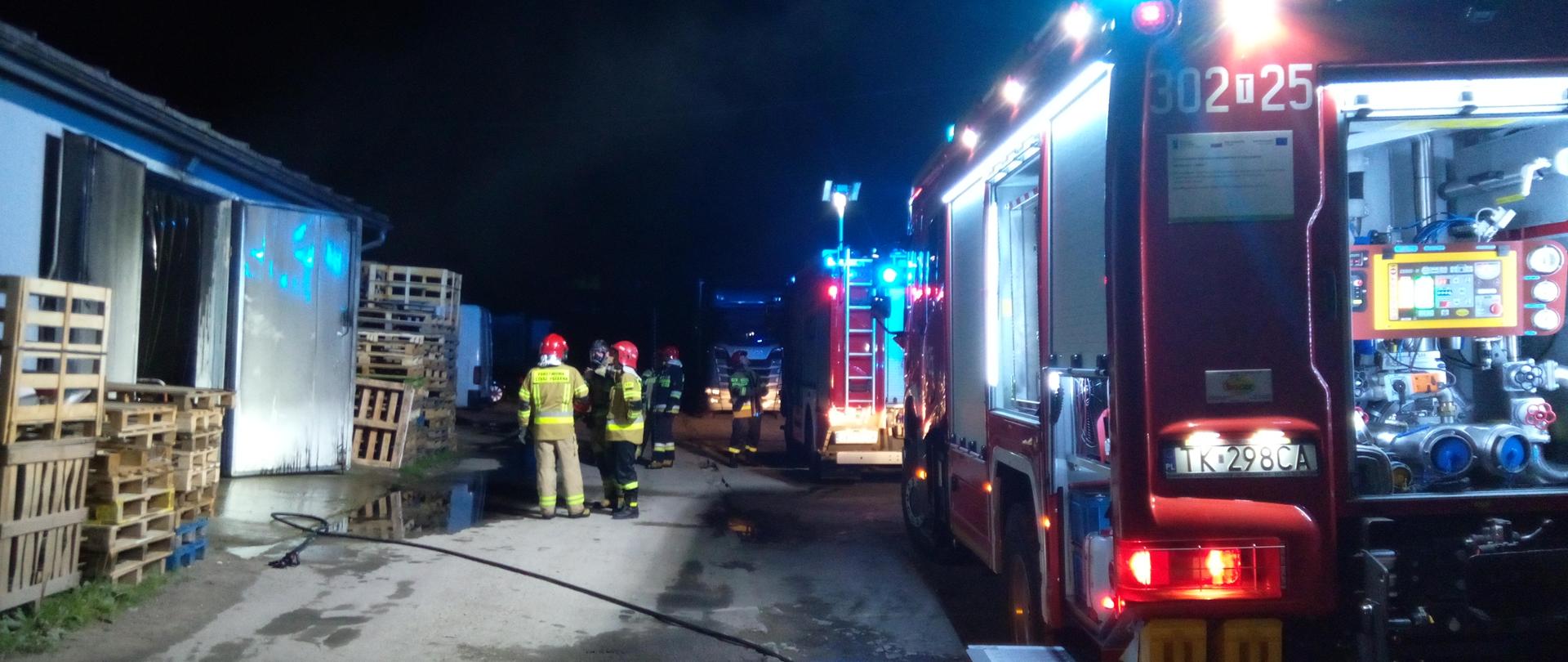 Na zdjęciu widać z prawej strony 2 wozy strażackie a po lewej halę magazynową, w której wybuchł pożar. Widać także grupę strażaków.