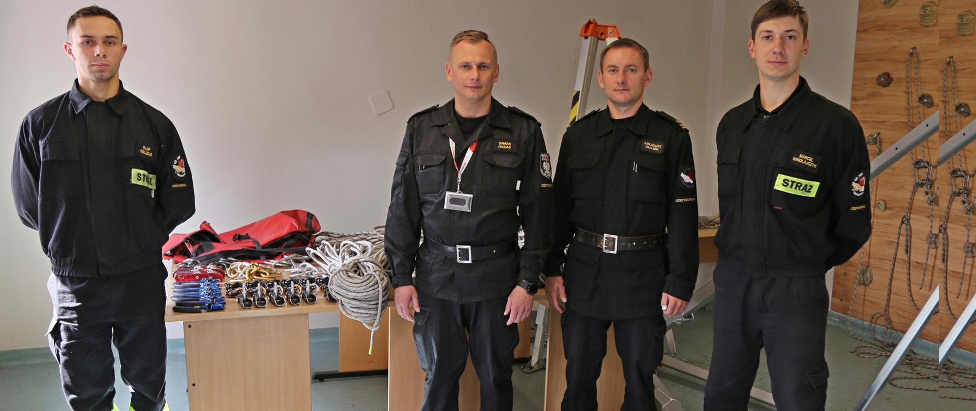 Na zdjęciu Zastępca Komendanta Centralnej Szkoły PSP w towarzystwie trach strażaków. Za nimi na biurkach widoczny przekazywany sprzęt ratownictwa wysokościowego