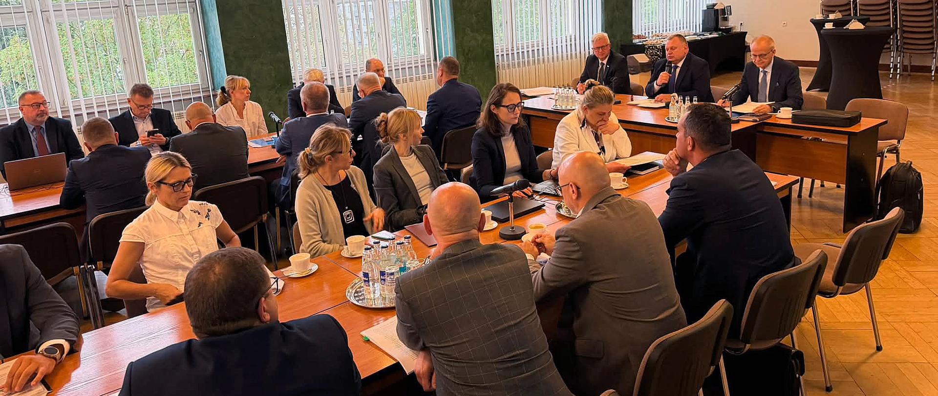 Grupa osób siedzi przy dwóch podłużnych stołach
