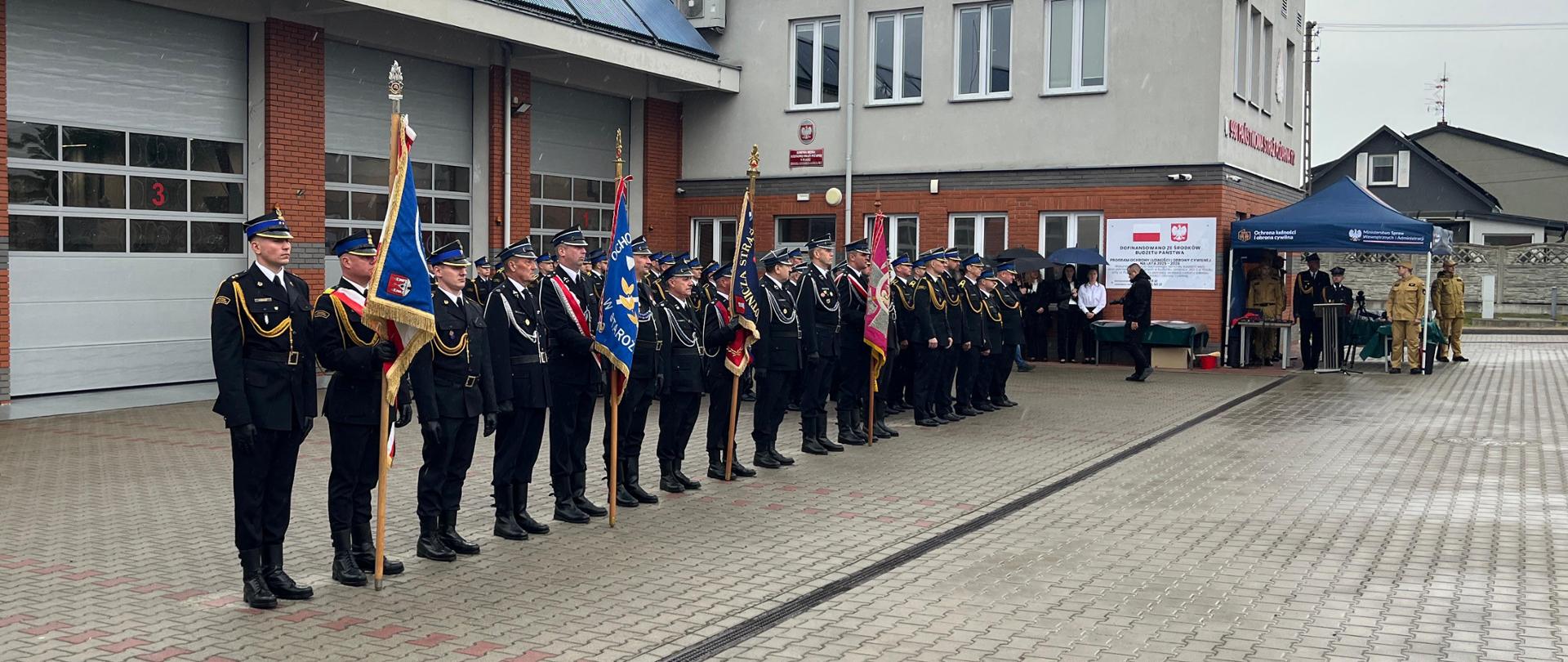 Strażacy w galowych mundurach stoją w szeregu podczas apelu z okazji Dnia Strażaka. W tle znajdują się namiot OLiOC oraz budynek jednostki.