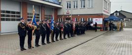 Strażacy w galowych mundurach stoją w szeregu podczas apelu z okazji Dnia Strażaka. W tle znajdują się namiot OLiOC oraz budynek jednostki.