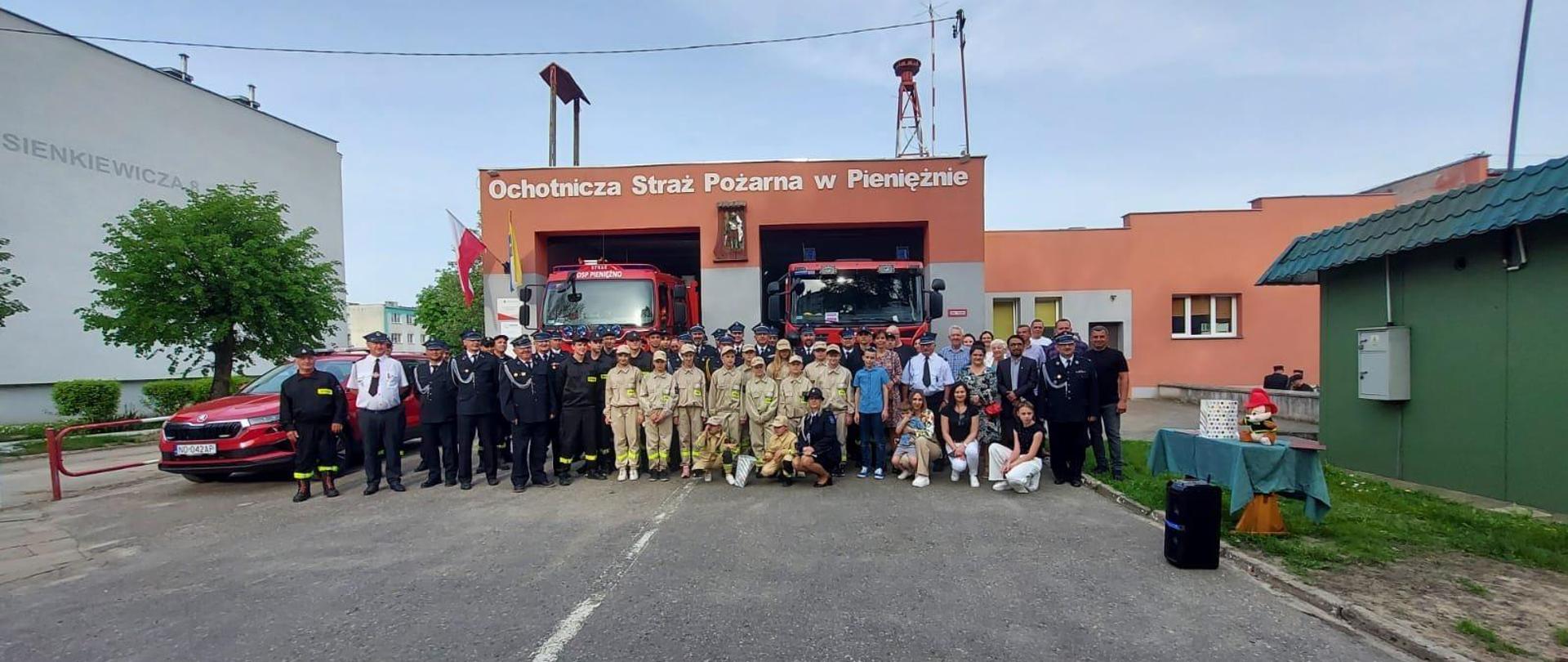 Przed remizą OSP do zdjęcia pozuje duża grupa strażaków ochotników w mundurach. W tle samochody strażackie.